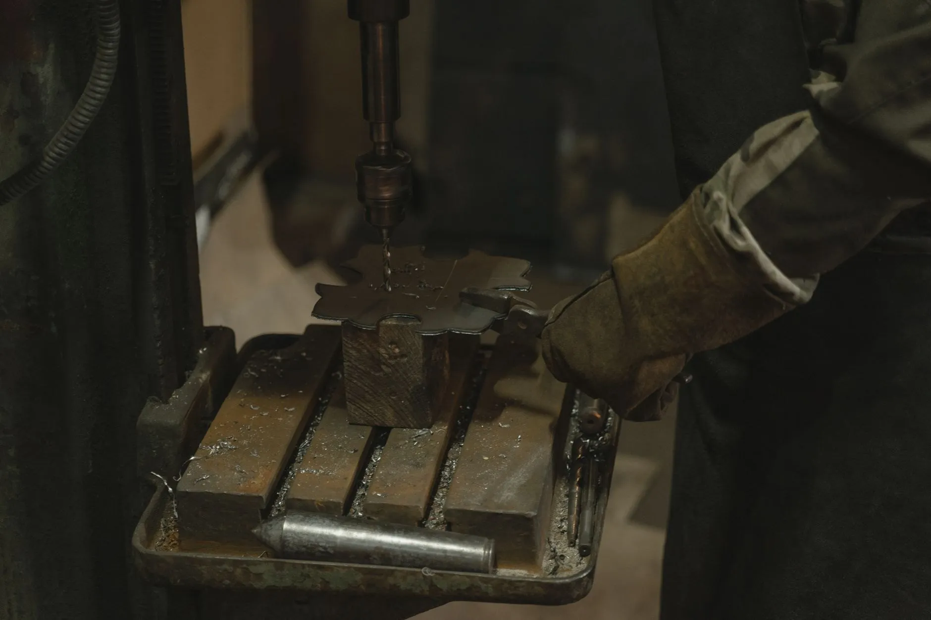 A gloved hand holding a steel part under a drill press