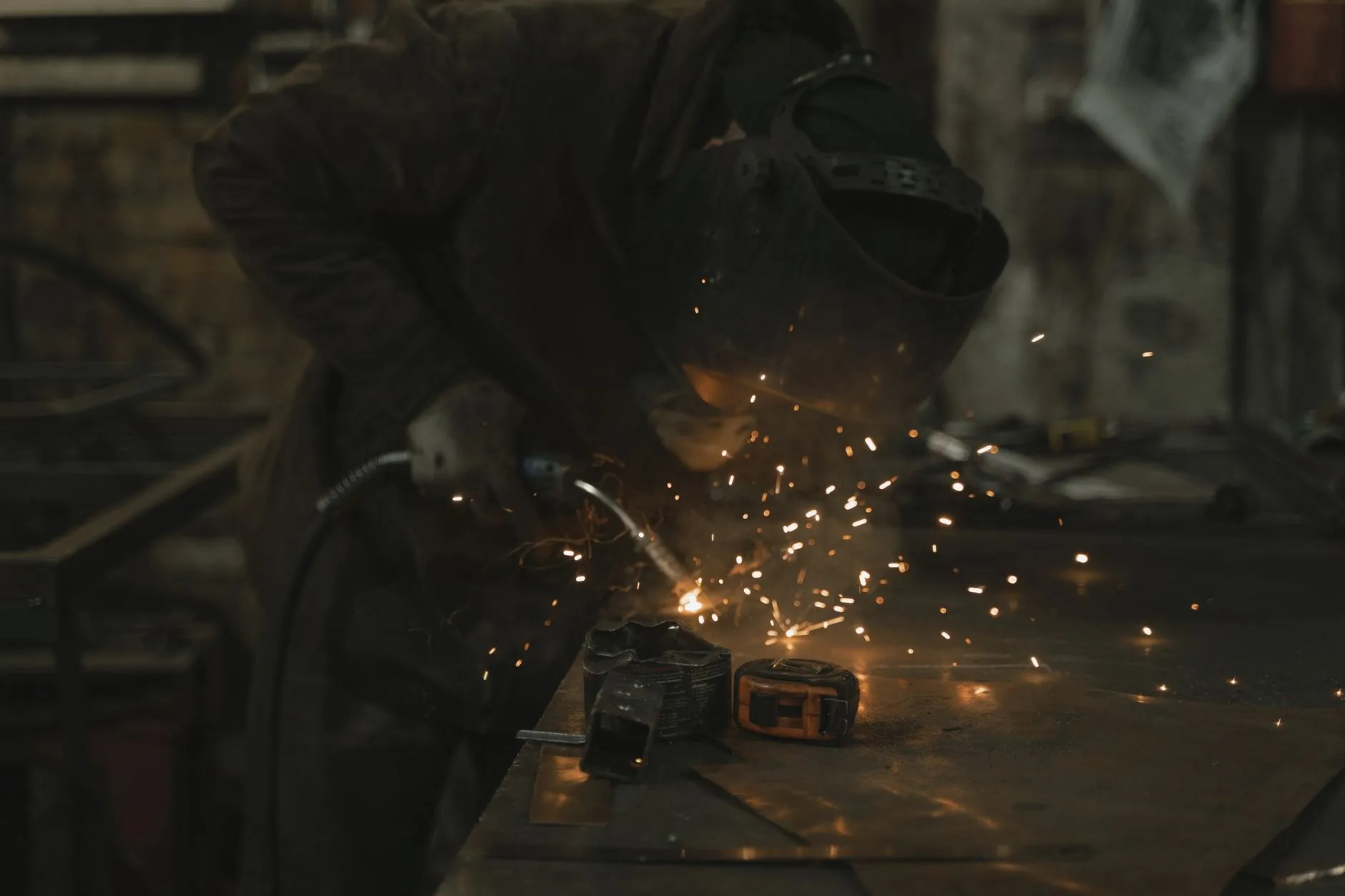A welder bent over a bench in a dim bay, sparks lifting off the joint