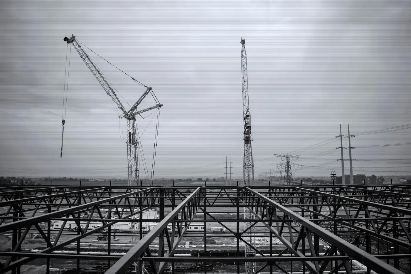 A steel roof frame under grey sky with two cranes behind it