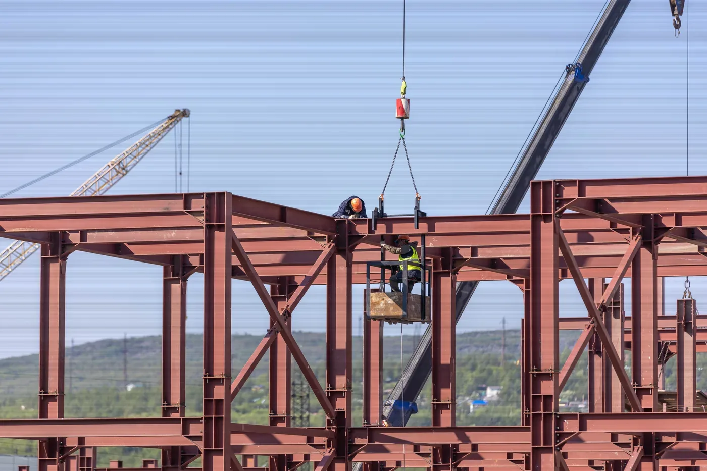 A steel frame being erected, a beam hanging from a crane