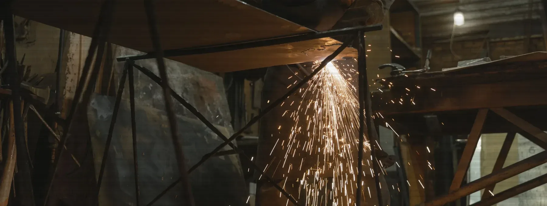 A fabricator dressing a weld on a steel frame, sparks falling to the shop floor